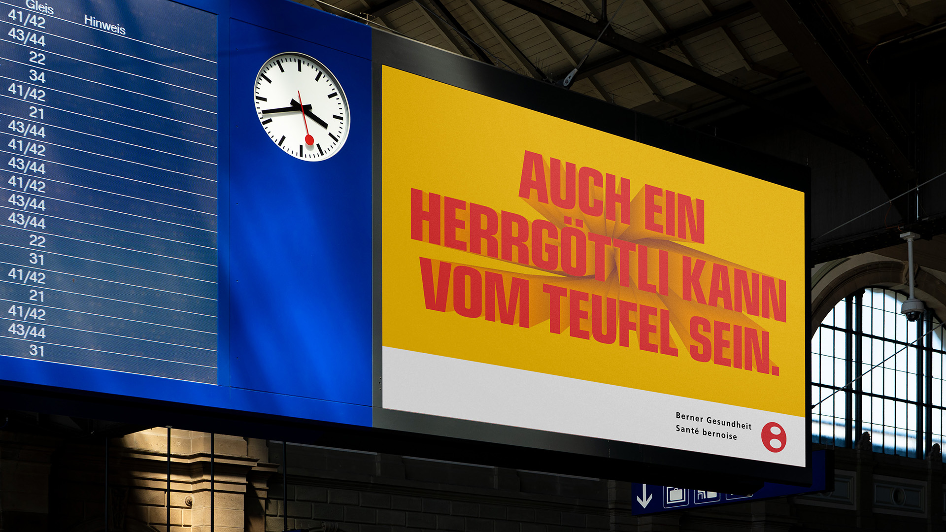 Digitale Bahnhofsanzeige mit gelbem Plakat und roter Schrift: „Auch ein Herrgöttli kann vom Teufel sein“, daneben eine Bahnhofsuhr.