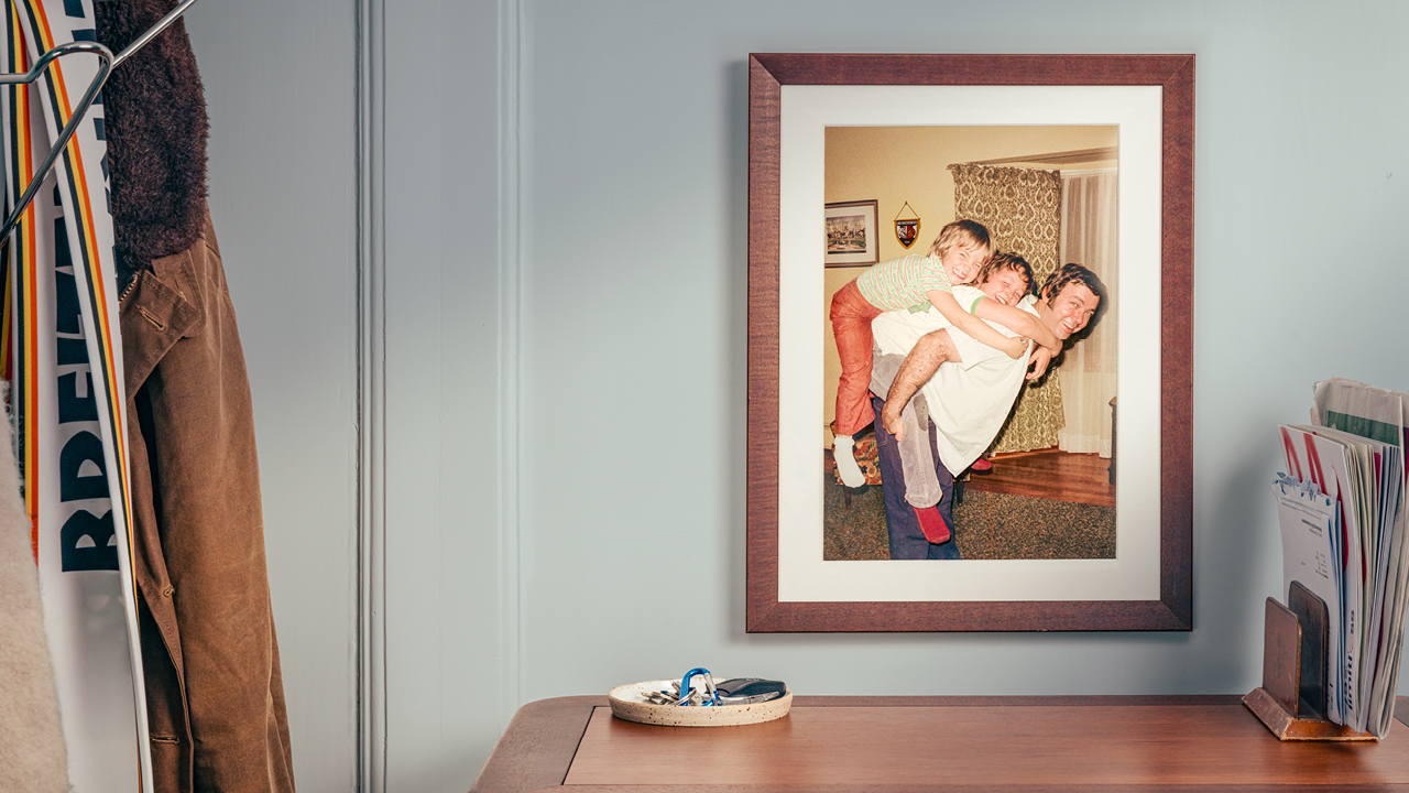 Gerahmtes Familienfoto an der Wand mit drei lachenden Personen, zwei davon auf dem Rücken des dritten, im Vordergrund eine Ablage für Schlüssel.