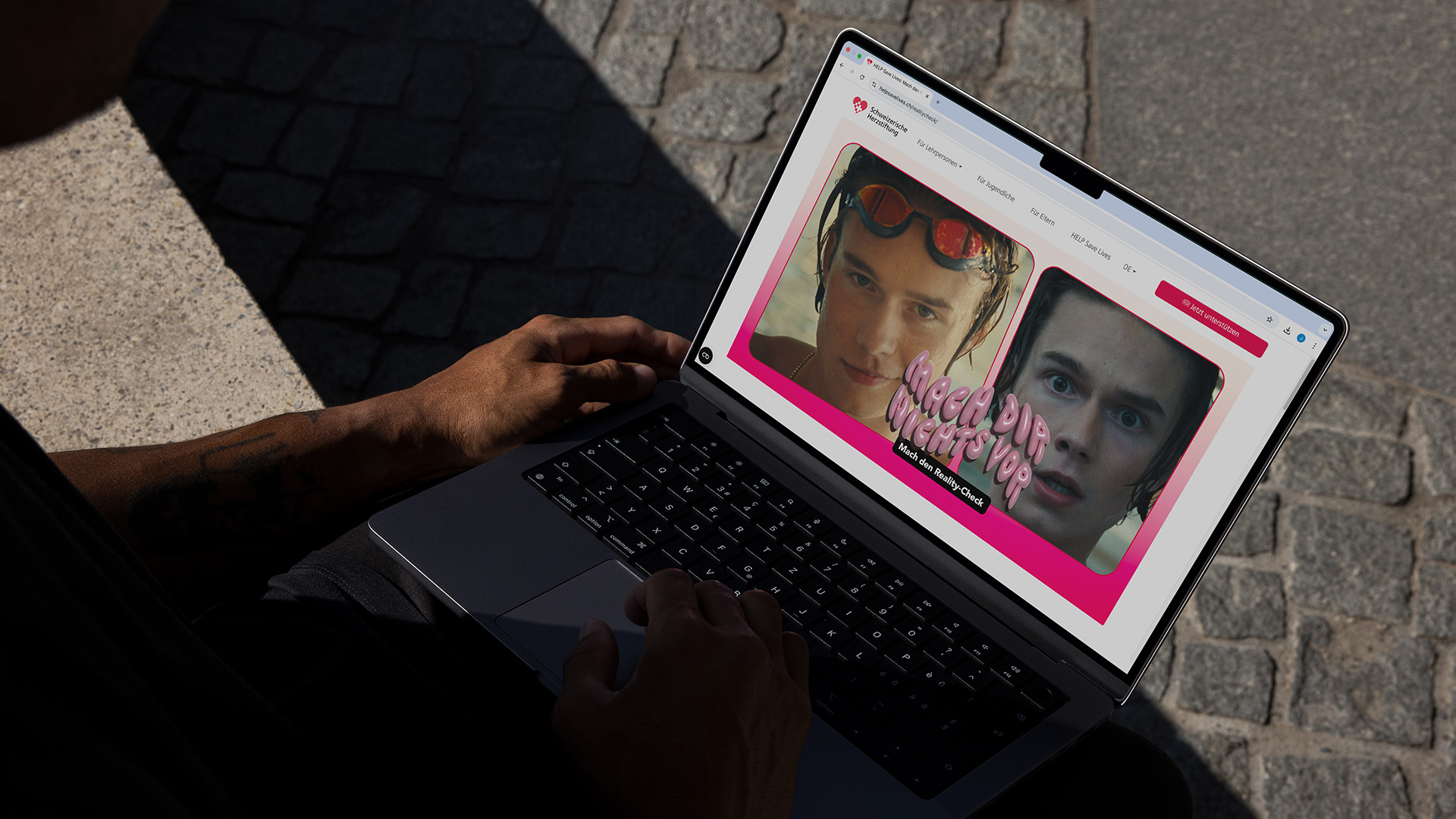 Person sitzt im Schatten mit einem Laptop auf dem Schoss, auf dem eine Website mit zwei Gesichtern und dem Text „Mach dir nichts vor“ angezeigt wird.