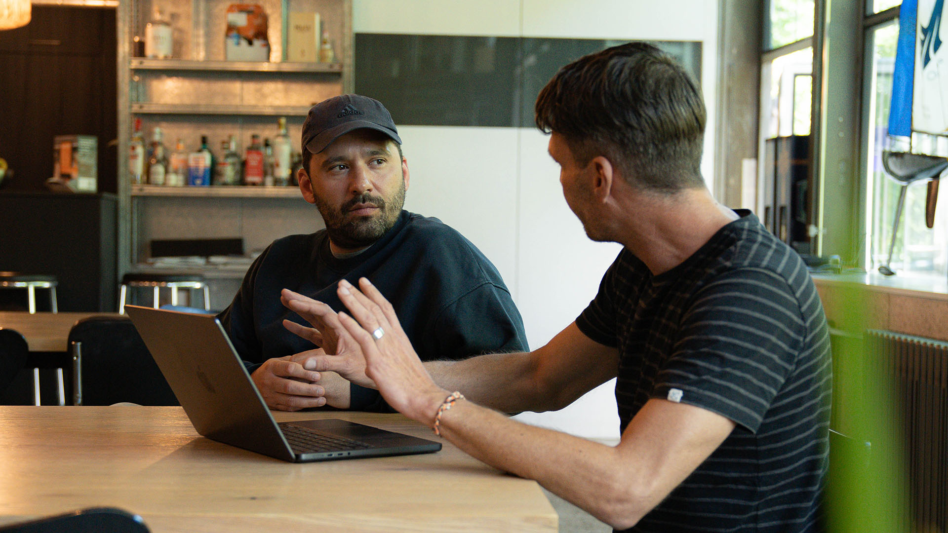 Zwei Männer sitzen an einem Tisch mit einem Laptop und unterhalten sich. im Hintergrund ein Regal mit Getränken.