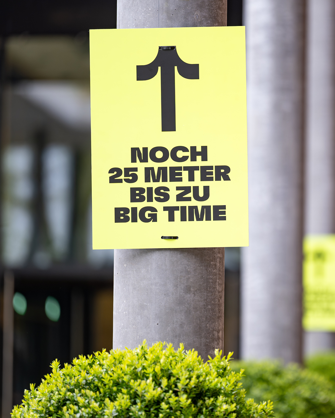 Ein gelbes Schild mit schwarzem Pfeil nach oben und der Aufschrift „NOCH 25 METER BIS ZU BIG TIME“ ist an einem Pfeiler befestigt, im Vordergrund grüne Büsche.
